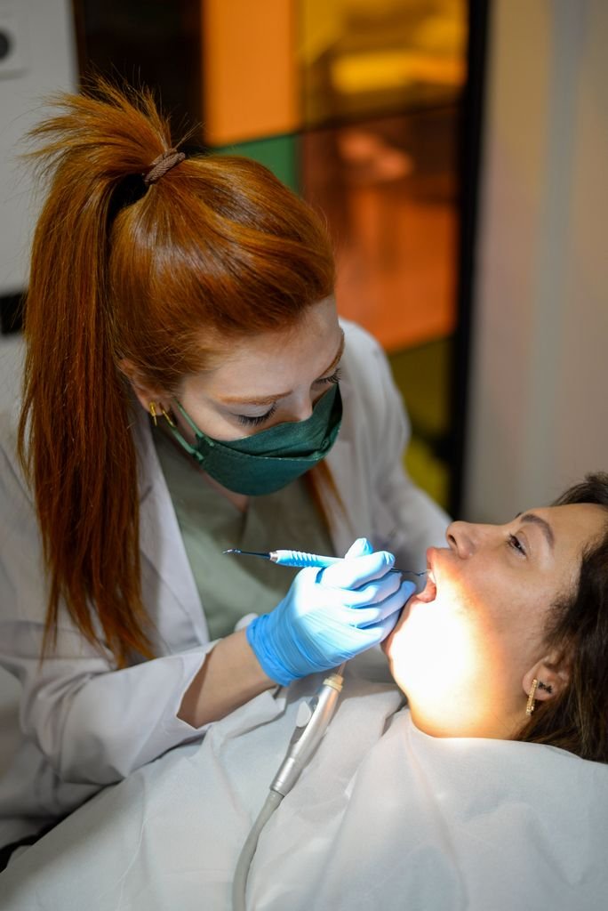 A female dentist in a mask and gloves examines a patient indoors under bright light.