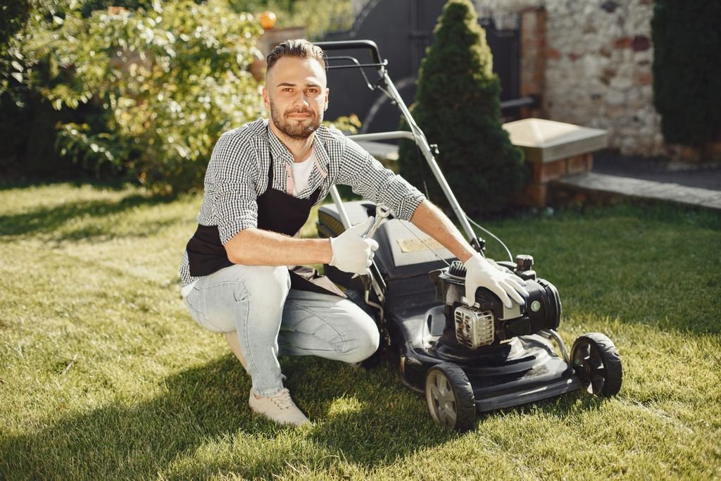 Adult man wearing apron and gloves maintains a lawn mower on a sunny day.